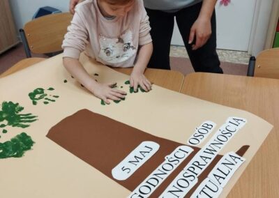 Dziecko robi zielone odbicia dłoni na plakacie o Dniu Godności Osób z Niepełnosprawnością Intelektualną.