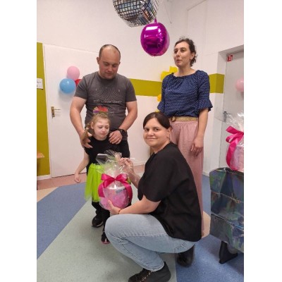 Mężczyzna z dziewczynką, kobieta klęcząca trzyma prezent, druga kobieta stoi w pomieszczeniu z dekoracjami.