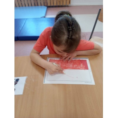 Dziewczynka koloruje na kartce papieru czerwoną kredką siedząc przy stoliku.