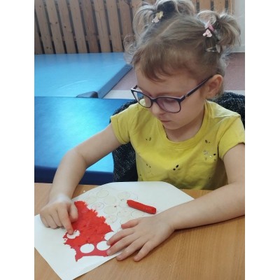 Dziewczynka z okularami nakleja czerwoną plastelinę na kartkę z narysowanymi kółkami.