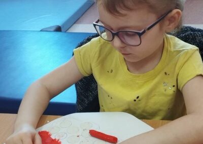 Dziewczynka z okularami nakleja czerwoną plastelinę na kartkę z narysowanymi kółkami.