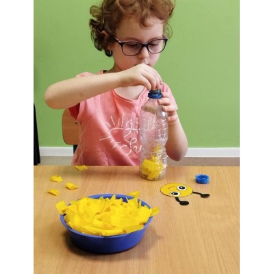 Dziewczynka wkłada żółte kawałki papieru do plastikowej butelki przy stole z miską i papierowym elementem.