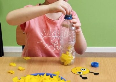 Dziewczynka wkłada żółte kawałki papieru do plastikowej butelki przy stole z miską i papierowym elementem.