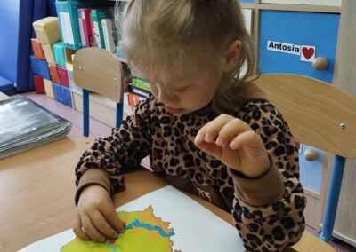 Dziewczynka siedzi przy stoliku i wskazuje palcem kolorową mapę na kartce papieru.