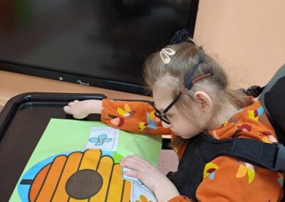 Dziewczynka na wózku inwalidzkim układa obrazki kwiatów na stole przed telewizorem.