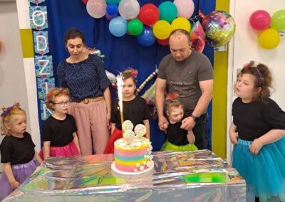 Grupa dzieci i dwoje dorosłych przy stole z urodzinowym tortem i balonami w tle.