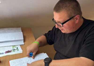 Mężczyzna z krótkimi włosami i okularami rysuje przy stole z otwartymi książkami i zeszytami.