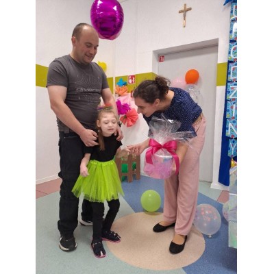 Mężczyzna trzyma dziewczynkę w zielonej spódniczce, kobieta podaje jej prezent na przyjęciu.