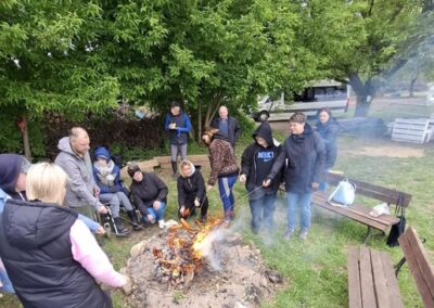 Ludzie wokół ogniska na trawie w parku, niektórzy trzymają patyki nad ogniem.