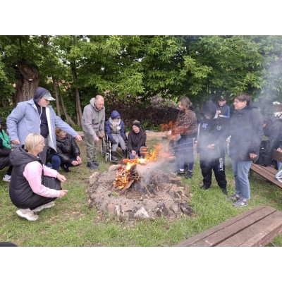 Grupa osób piekąca kiełbaski na ognisku na zewnątrz w otoczeniu drzew i ławek.