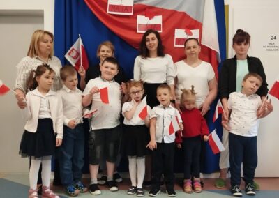 Grupa dzieci i dorosłych z małymi flagami Polski przed dekoracją z flagą i godłem na 3 maja.