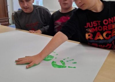 Trzech chłopców przy stole, jeden odciska dłoń z zieloną farbą na dużym arkuszu papieru.