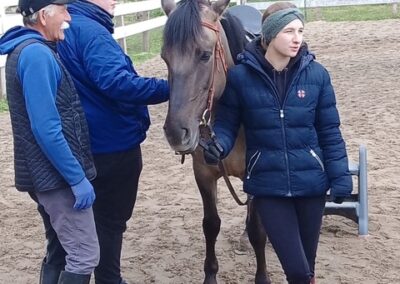 Trzy osoby stoją przy koniu na piaskowym placu w otoczeniu ogrodzenia i zieleni.