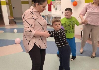 Dwie kobiety i dwóch chłopców trzymają się za ręce w sali z balonami i tablicą ogłoszeń.