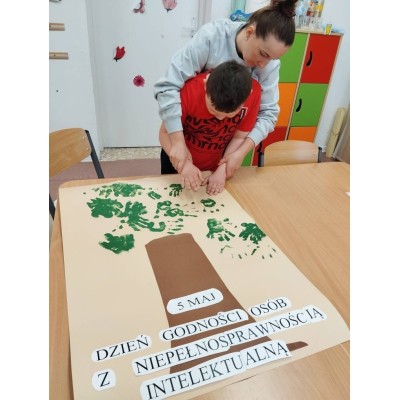 Kobieta pomaga chłopcu w tworzeniu plakatu o Dniu Godności Osób z Niepełnosprawnością Intelektualną.