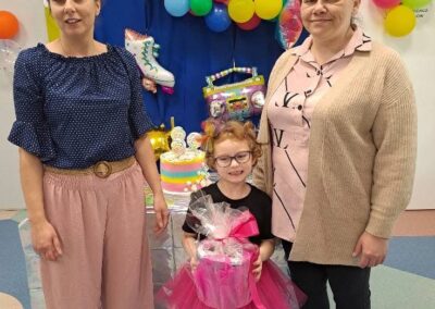 dwie kobiety i dziewczynka w różowej spódniczce z prezentem na tle balonów i urodzinowych dekoracji