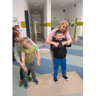 Dwie kobiety pomagają dwóm chłopcom w ćwiczeniach ruchowych na kolorowej podłodze w jasnym pomieszczeniu.