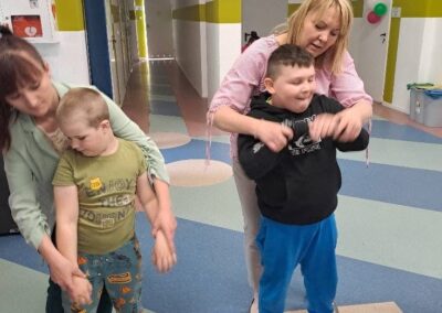 Dwie kobiety pomagają dwóm chłopcom w ćwiczeniach ruchowych na kolorowej podłodze w jasnym pomieszczeniu.