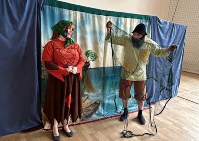 Dwóch aktorów w ludowych strojach na scenie z dekoracją nadbrzeża i boisko szkolne w tle.