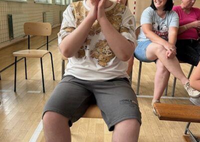 Chłopiec w czapce siedzi na krześle w sali gimnastycznej, w tle trzy kobiety na ławkach.