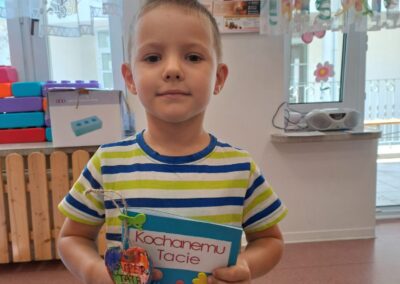 Chłopiec trzyma kartkę Kochanemu Taci i ozdobiony breloczek w przedszkolu.