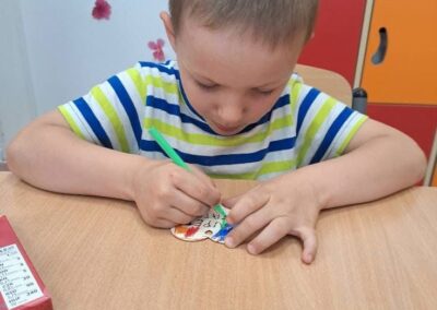 Chłopiec koloruje papierowy kształt zielonym flamastrem siedząc przy stoliku w przedszkolu.