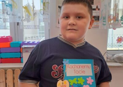 Chłopiec pokazuje laurkę z napisem Kochanemu Tacie ozdobioną kolorowymi naklejkami, w tle kalendarz.