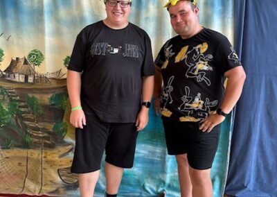 Dwóch mężczyzn w czarnych ubraniach i żółtych nakryciach głowy na tle malowanego wiejskiego pejzażu.