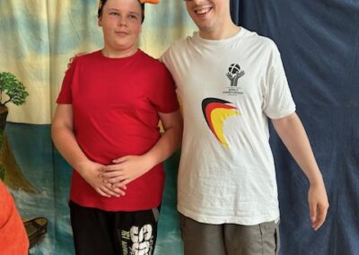 Dwaj mężczyźni w krótkich spodenkach i kolorowych czapkach stoją przy zasłonie w sali sportowej.