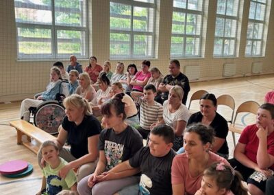 Grupa dzieci i dorosłych siedzi na ławkach w sali gimnastycznej z dużymi oknami.