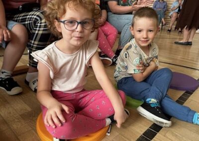 Dzieci i dorośli siedzą na podłodze sali gimnastycznej na kolorowych poduszkach.