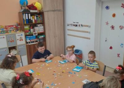 Dzieci siedzą przy stole w klasie, wykonując prace plastyczne z kolorowymi elementami.