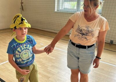 Dziecko w czapce z postacią z kreskówki trzyma dłoń dorosłej kobiety na sali gimnastycznej.