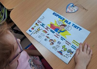 Dziecko siedzi przy stoliku i trzyma kartkę z kuponami dla taty oraz serduszko z napisem.