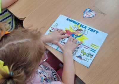 Dziecko siedzi przy stole i trzyma kupon zatytułowany Kupony dla Taty z kolorowymi elementami.