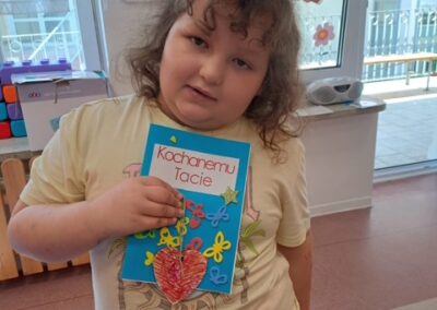 Dziewczynka pokazuje kartkę dla taty ozdobioną sercem, motylkami i gwiazdką.