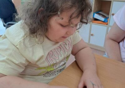 Dziewczynka koloruje różowym flamastrem owalny przedmiot siedząc przy stole w pomieszczeniu.