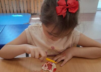 Dziewczynka z czerwonymi kokardami koloruje papierowe serce żółtym mazakiem przy stole.