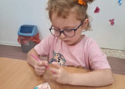 Dziewczynka z okularami maluje wycinankę serca różowym pisakiem przy stole.