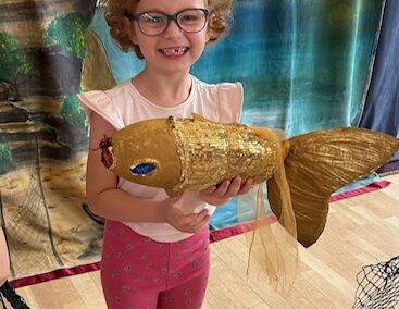 Dziewczynka w okularach trzyma dużą złotą rybę z papieru, stoi na tle nadmorskiej dekoracji.