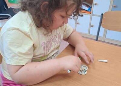 Dziewczynka rysuje przy stoliku, na tle szafek z pojemnikami i materiałami edukacyjnymi.