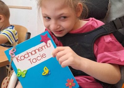 Dziewczynka na wózku trzyma kartkę Kochanemu Tacie ozdobioną motylkami i kwiatami.