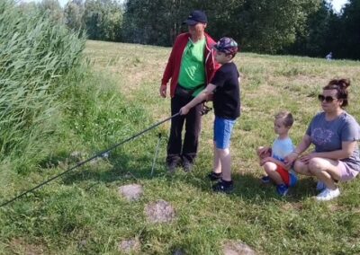 Cztery osoby nad wodą na trawie, dwoje dzieci łowi ryby, dwoje dorosłych obserwuje.