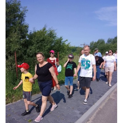 Grupa dorosłych i dzieci spaceruje chodnikiem w słoneczny dzień obok zieleni i drzew.