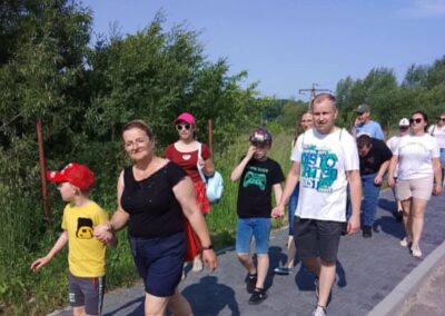 Grupa dorosłych i dzieci spaceruje chodnikiem w słoneczny dzień obok zieleni i drzew.