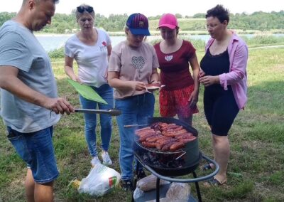 Pięć osób grilluje kiełbaski na grillu na trawie nad jeziorem w słoneczny dzień.