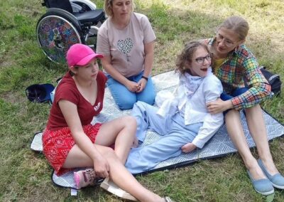 Cztery osoby siedzą na kocu na trawie w parku obok kolorowego wózka inwalidzkiego.