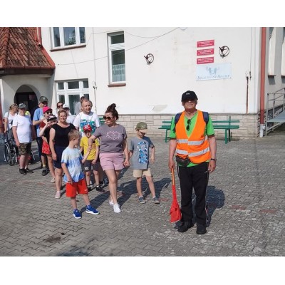Grupa dorosłych i dzieci stoi na placu z kostki brukowej przed budynkiem, mężczyzna w kamizelce odblaskowej trzyma czerwoną miotłę.