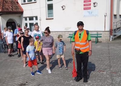 Grupa dorosłych i dzieci stoi na placu z kostki brukowej przed budynkiem, mężczyzna w kamizelce odblaskowej trzyma czerwoną miotłę.