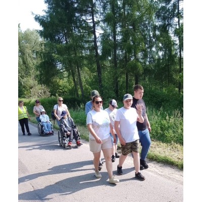 Grupa osób spacerująca po leśnej drodze, w tym dwie osoby na wózkach inwalidzkich.